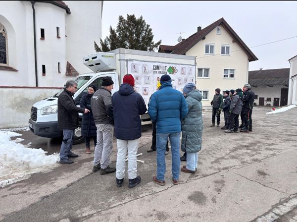 Eine Gruppe von Menschen steht im Winter vor dem weißen Werbe-Lkw der COMPAKT-Liste Karlskron in Adelshausen. Im Hintergrund ist eine Kirche und ein Wohnhaus zu sehen.