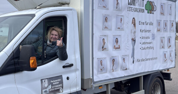 Eine lächelnde Frau am Steuer eines weißen Transporters mit einer Werbeplane der „COMPAKT - Liste Karlskron“. Sie zeigt den Daumen nach oben. Auf der Plane sind Team-Fotos und politische Slogans zu sehen.