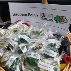 Ein Weidenkorb voller handgemachter, grün-marmorierter Seifenstücke, die einzeln verpackt sind. Jede Seife trägt eine Banderole mit der Aufschrift „Saubere Politik“ und dem Logo der „COMPAKT - Liste Karlskron“.