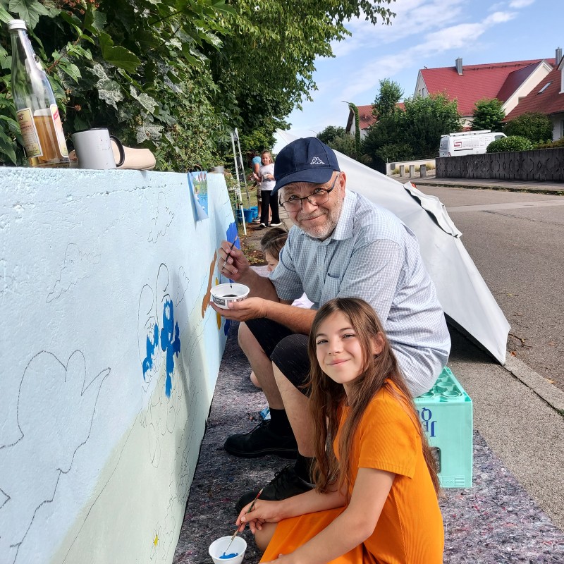 Ein älterer Mann mit Mütze und Brille und ein junges Mädchen mit orangefarbenem Kleid malen gemeinsam ein Schlümpfe-Wandbild an einer Wand im Freien.