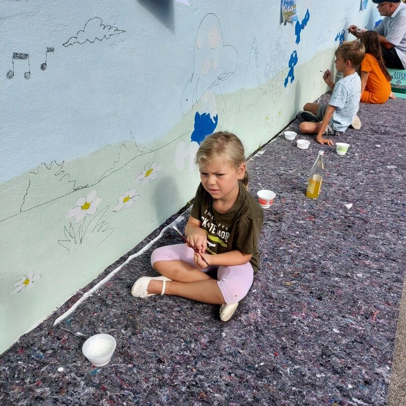 Mehrere Kinder sitzen auf Malervlies entlang einer niedrigen Betonwand und zeichnen sowie malen mit Pinseln an einem Schlümpfe-Wandbild.