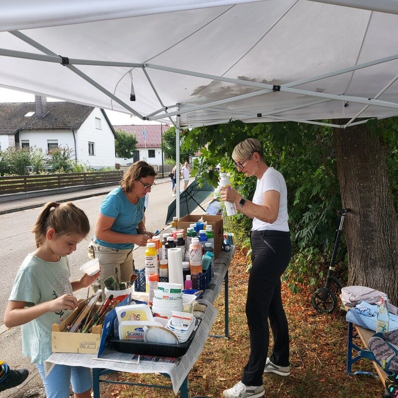 Zwei Frauen und ein Mädchen stehen unter einem weißen Faltpavillon an einem Tisch voller Farbdosen, Becher und Pinsel zur Vorbereitung der Malaktion.