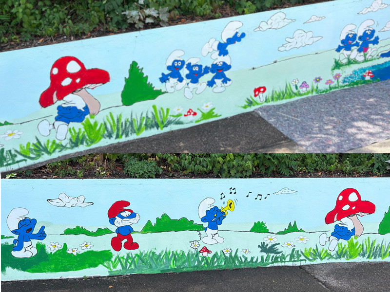 Bunt bemaltes Wandgemälde mit Schlümpfen in einer Wiesenlandschaft, darunter Papa Schlumpf, ein Trompete spielender Schlumpf und Figuren mit Fliegenpilzen.