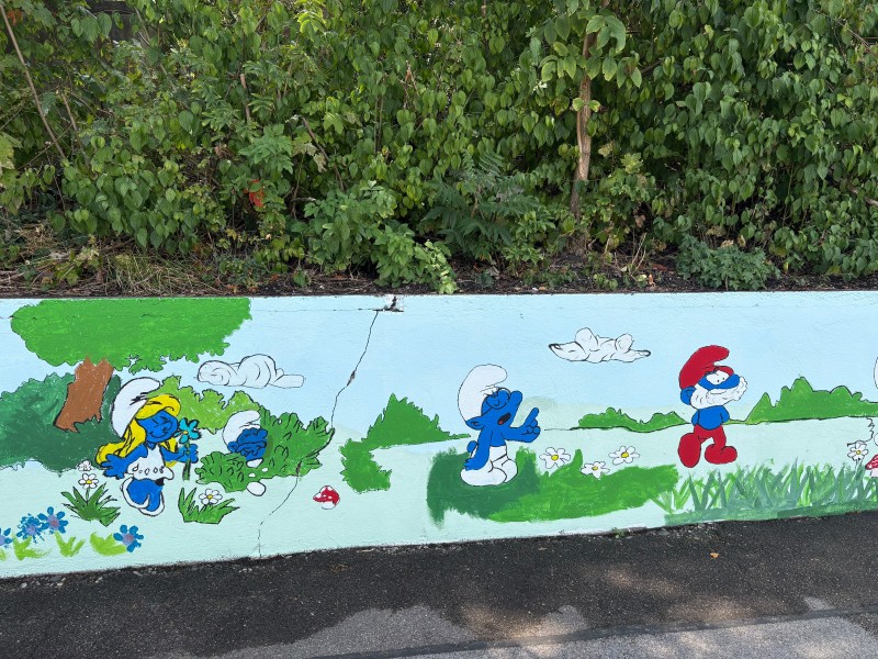 Handgemaltes Wandbild an einer Außenmauer mit den Figuren Schlumpfine, einem betenden Schlumpf und Papa Schlumpf in einer grünen Wiesenlandschaft unter blauem Himmel.