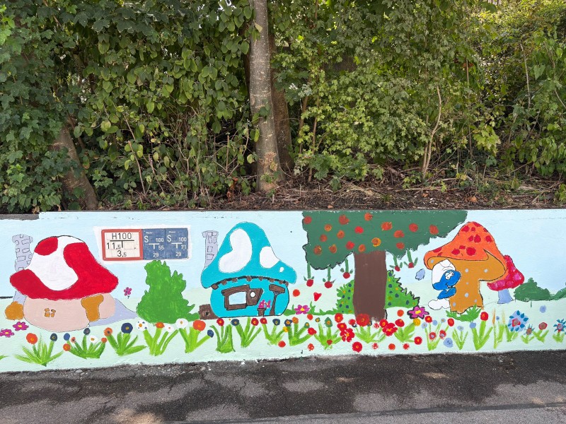 Ein handgemaltes Wandbild auf einer niedrigen Betonmauer, das eine Szene aus Schlumpfhausen zeigt, mit Pilzhäusern, einem Apfelbaum und einem Schlumpf. Im oberen Teil sind Schilder mit technischen Beschriftungen wie "H100" und "S100" zu sehen.