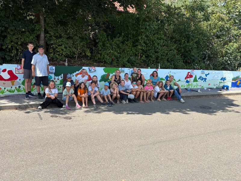 Eine große Gruppe von Kindern und Erwachsenen sitzt und steht fröhlich vor einer frisch bemalten Wand mit bunten Schlümpfe-Motiven in Karlskron.