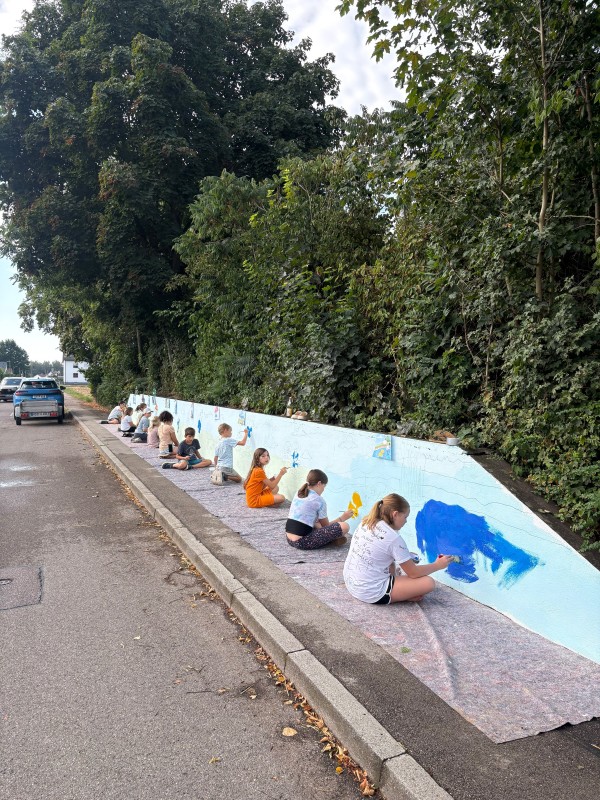 Eine Reihe von Kindern sitzt auf ausgelegtem Malervlies am Straßenrand vor einer langen, hellblau grundierten Betonwand und malt an einem großen Wandbild.