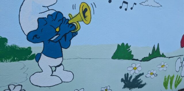 Ein Wandgemälde, das einen Schlumpf zeigt, der auf einer Trompete spielt, während Musiknoten in der Luft schweben, auf einer bemalten Wand mit Gänseblümchen und Pilzen.